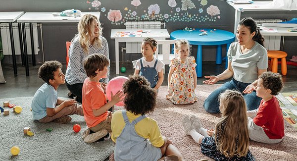 Zwei Erzieherinnen sitzen mit vielen kleinen Kindern in einem Sitzkreis