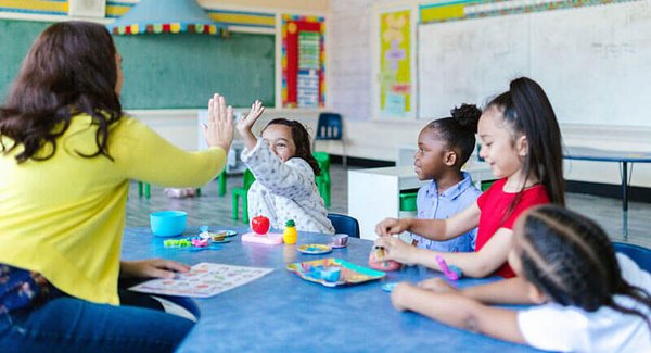 Lehrerin sitzt mit vier Schülerinnen am Tisch, eine davon gibt sie ein High five