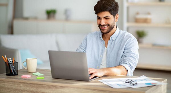 Ein junger Mann sitzt mit Schreibutensilien Zuhause vor einem Laptop und lächelt