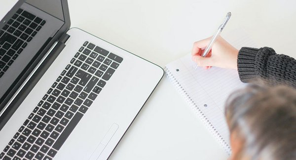 Aus der Vogelperspektive ist links ein Laptop und rechts eine Hand, die etwas auf einen Block schreibt, zu sehen