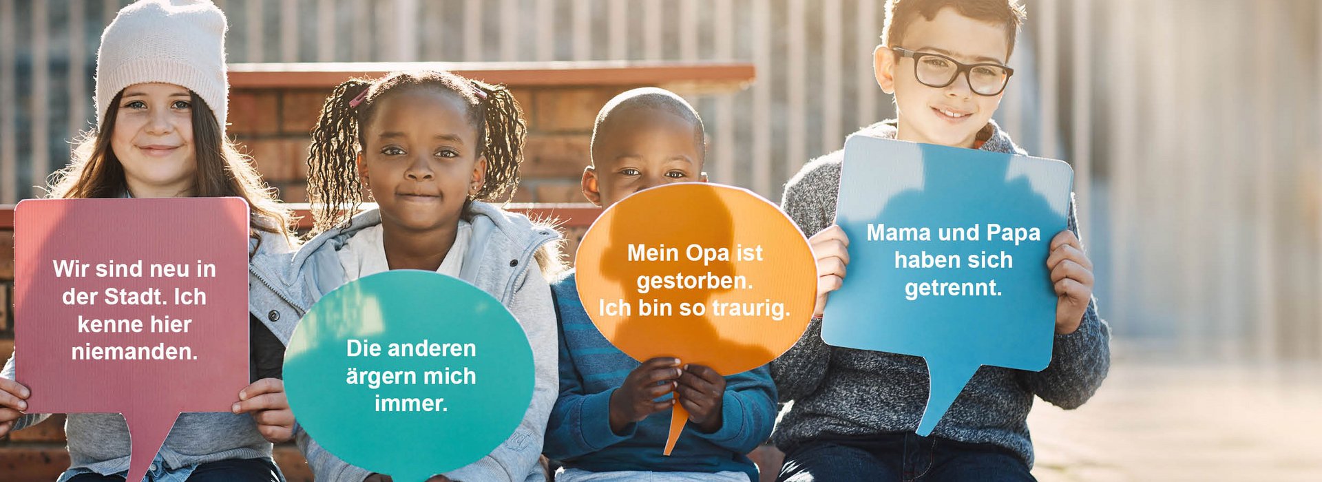 Vier Kinder sitzen auf einer Bank und halten bunte Sprechblasen in die Kamera