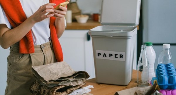 Eine junge Frau steht an einem Tisch, auf dem ein Mülleimer steht, und trennt Plastik- und Papiermüll.